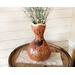 A curvy gourd vase, with a hummingbird on the front, sits on a whiskey barrel in an entryway corner. The vase holds branches with flowers. 